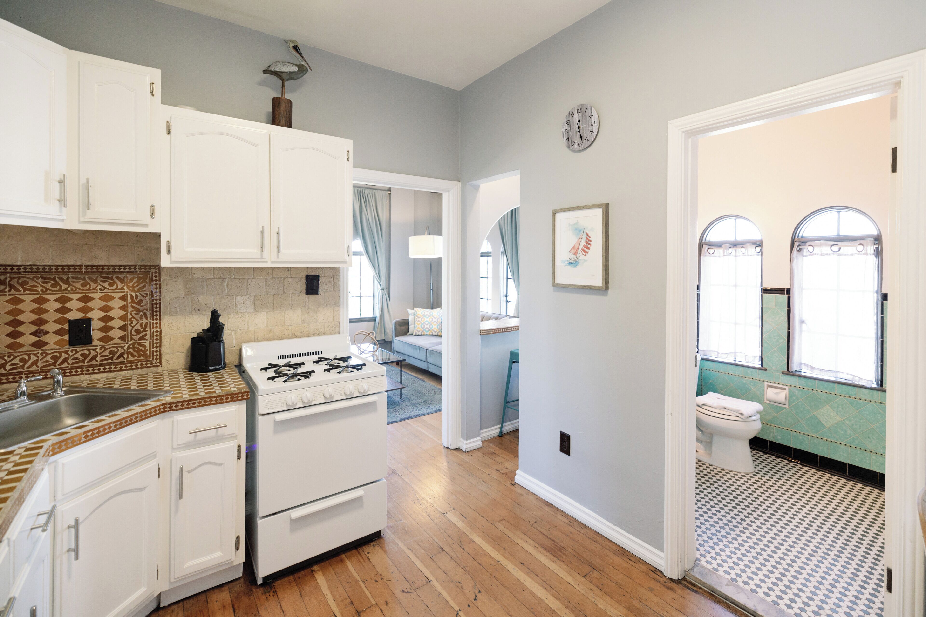 Une cuisine avec des meubles blancs, une cuisinière et un évier. Un couloir mène à une salle de bain équipée de toilettes et d’une fenêtre.