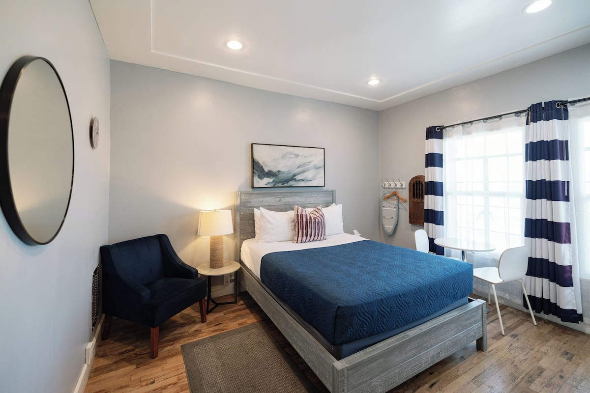 Une chambre à coucher comprenant un lit, une chaise, une table de chevet, une lampe, une horloge, un tableau et une fenêtre avec des rideaux.