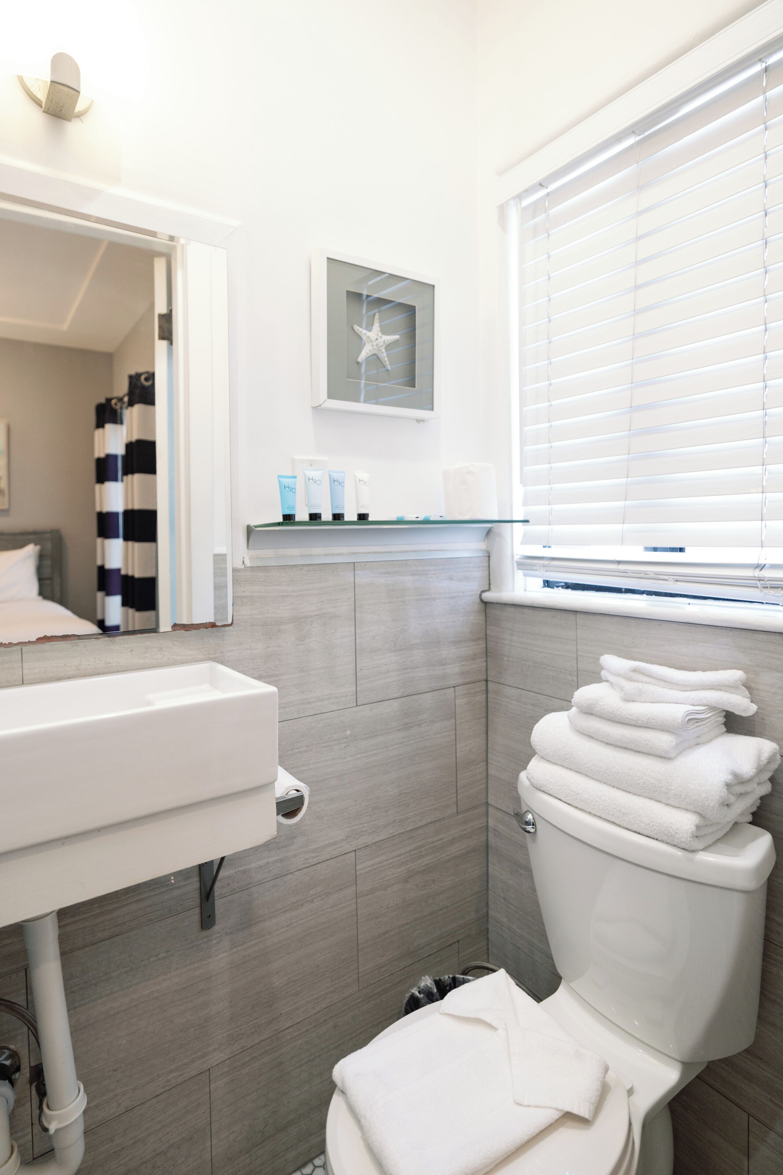 A modern bathroom with a white sink, a toilet with stacked towels, a starfish decoration, and a window with blinds.