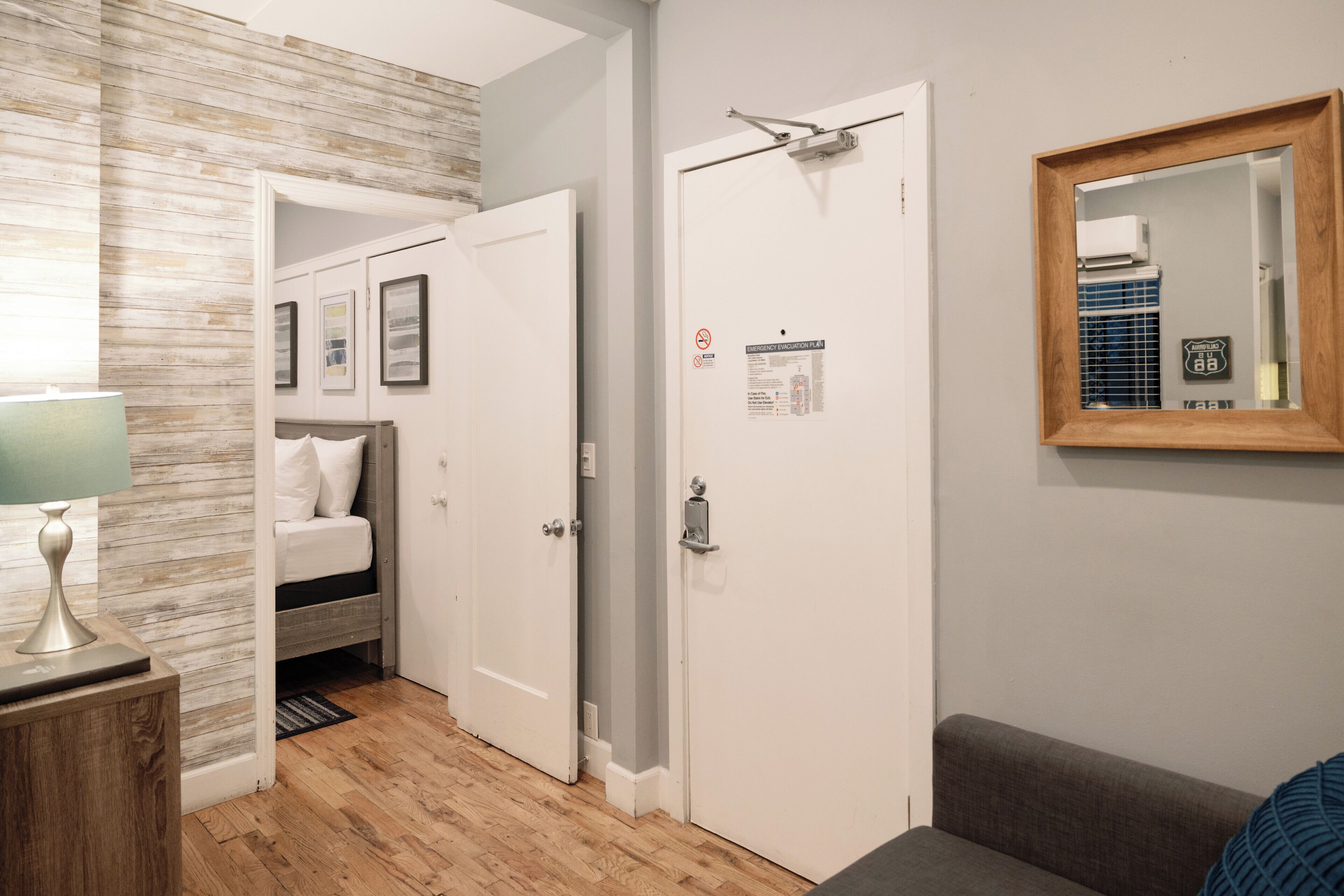 A hotel room with a wooden floor, a bed with white bedding, a bedside table with a lamp, a door with a mirror, and a wall-mounted thermostat.