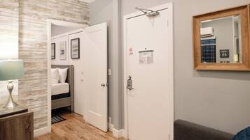 A hotel room with a wooden floor, a bed with white bedding, a bedside table with a lamp, a door with a mirror, and a wall-mounted thermostat.