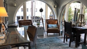 A spacious living room with a glass coffee table, two armchairs, a sofa, and a dining table. Large arched windows provide natural light and a view of the outside.
