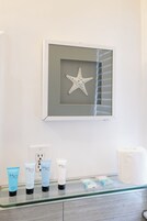 Un baño con una obra de arte de una estrella de mar enmarcada en la pared, un estante de vidrio con productos para el cuidado de la piel y un rollo de papel higiénico blanco.