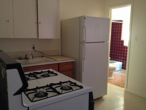 Una cocina con estufa a gas, alacenas blancas, un refrigerador y vista hacia un baño con pared de azulejos rojos.