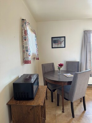 Un comedor con una mesa redonda, seis sillas, un microondas sobre un soporte de madera, un cuadro en la pared y una ventana con cortinas estampadas.