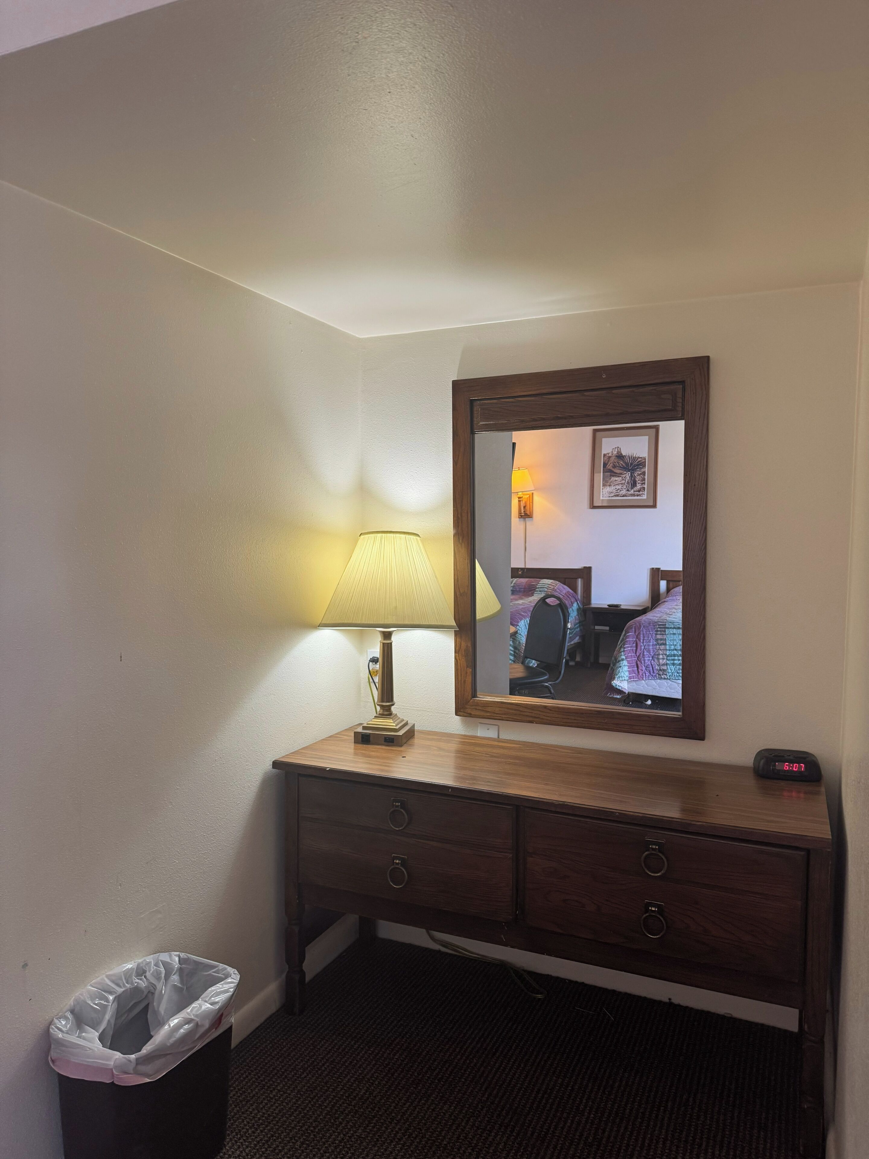 A hotel room with a wooden dresser, a mirror, a lamp, a trash bin, and a digital clock.