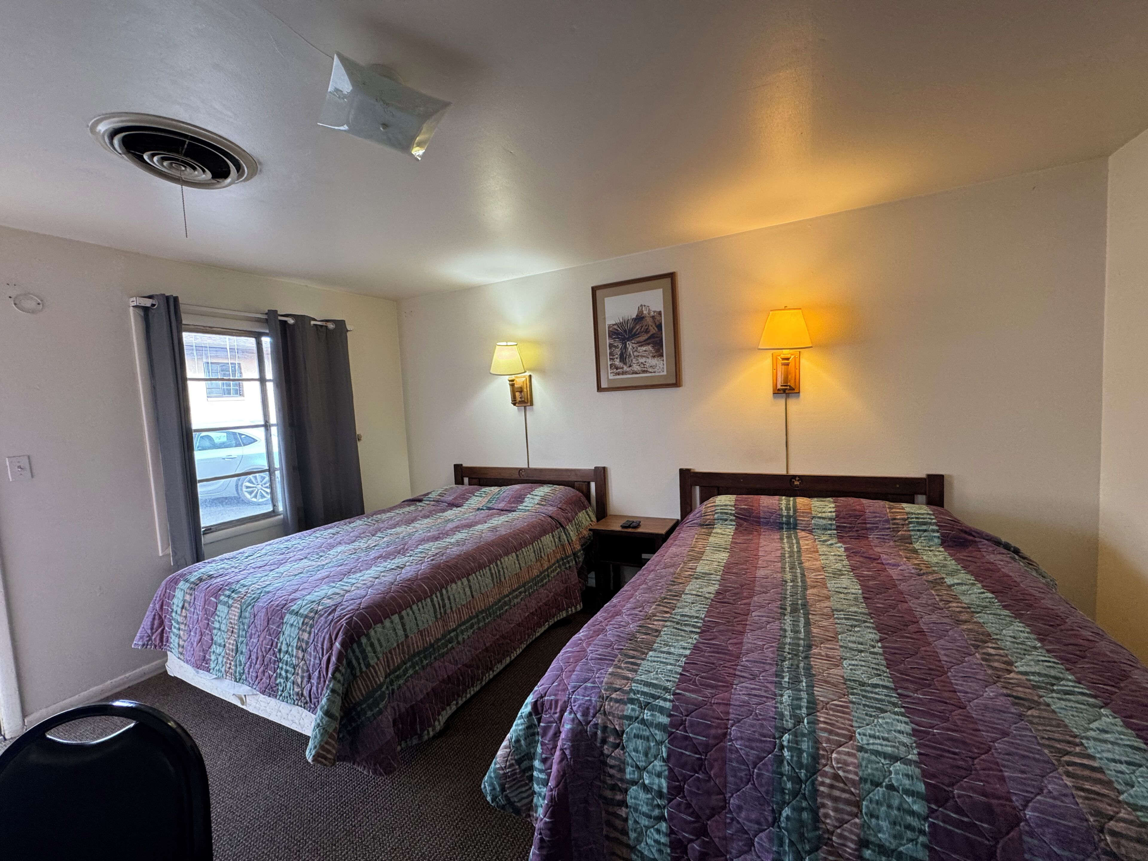 A hotel room with two beds, a window with curtains, a wall-mounted lamp, and a ceiling vent.