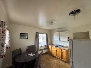 A compact kitchen with a dining table, a window with curtains, a white refrigerator, and a wall-mounted cabinet. - Cody Motel Rock Springs WY (Rock Springs)