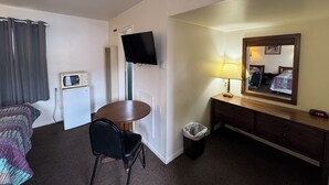 A hotel room with a bed, a desk, a chair, a television, a lamp, a mirror, and a trash bin.
