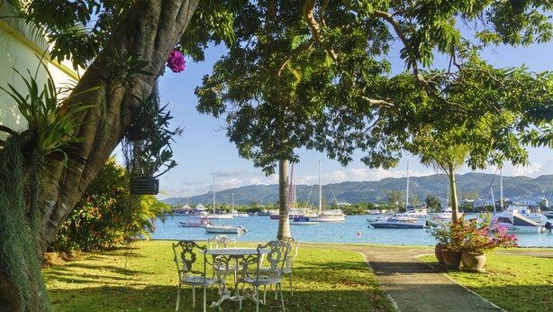 A grassy area with a pathway leading to a seating area with metal chairs and tables, overlooking a marina with boats. - Seawind On The Bay Beach Condo OLR (Montego Bay)