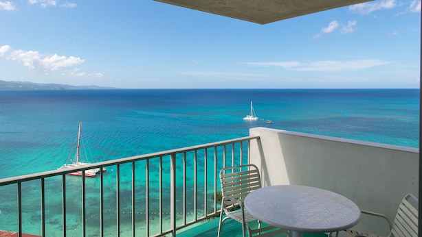 A balcony with a table and chairs overlooking turquoise waters and a sailboat. - Sunset Beach Studio At Montego Bay Club Resort (Montego Bay)