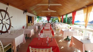 A restaurant with white tables and chairs, a large ship's wheel on the wall, and a view of the sea. - Sunset Beach Studio At Montego Bay Club Resort (Montego Bay)