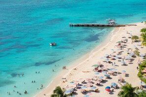 A beach with clear turquoise water, a pier, and numerous umbrellas and beach chairs. - Sundown Beach Studio At Montego Bay Club (Montego Bay)