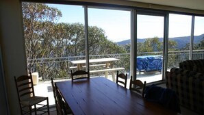 Un balcón con una mesa y sillas de madera, un sofá a cuadros y vistas a árboles y colinas.