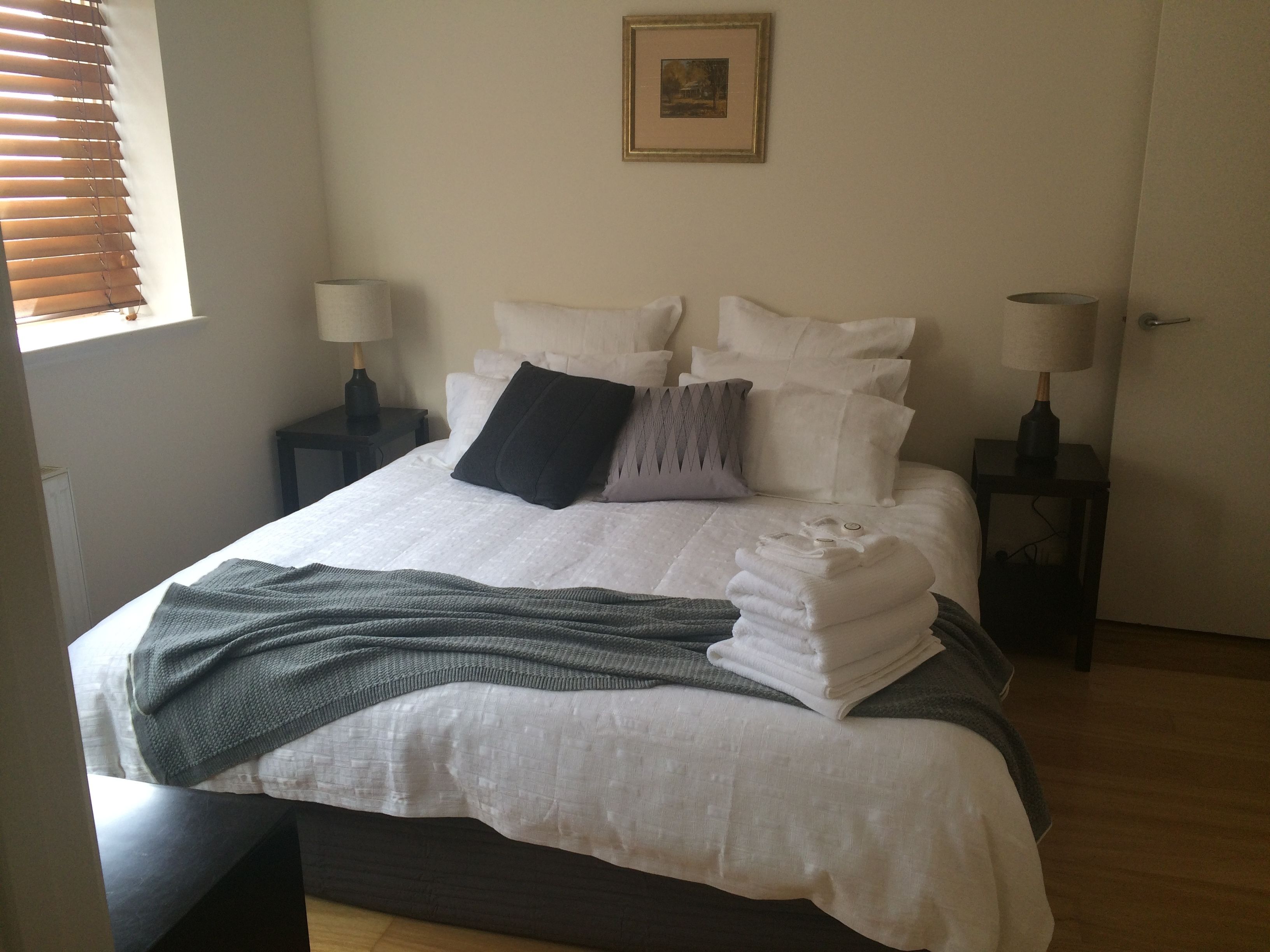 Un lit bien fait, avec du linge de lit blanc, une couverture foncée et des serviettes pliées. Il y a deux tables de chevet avec des lampes, un tableau encadré au mur et une fenêtre avec des stores.