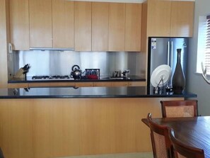 A kitchen with wooden cabinets, a stainless steel countertop, a refrigerator, and a dining table with chairs. - Mark II 4 West (Mount Buller)