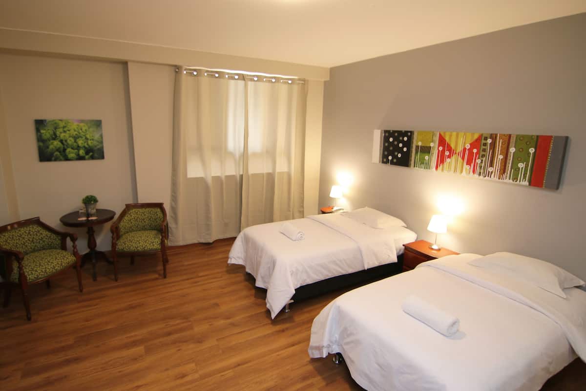 Una habitació d'hotel amb dos llits, una taula petita amb cadirees, un quadre a la paret i una finestra amb cortines.