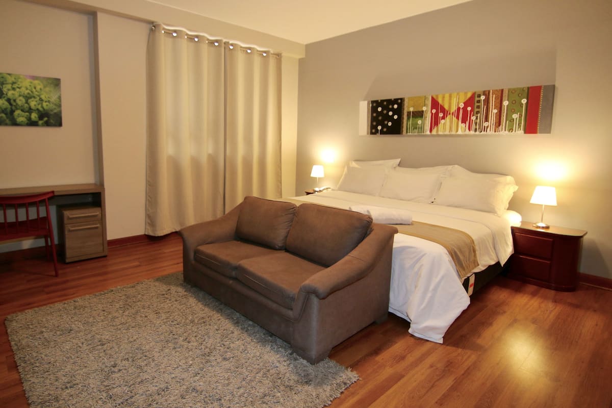 Una habitació d'hotel amb un llit, dues làmpades de nit, un sofà, una taula de treball i un quadre a la paret.