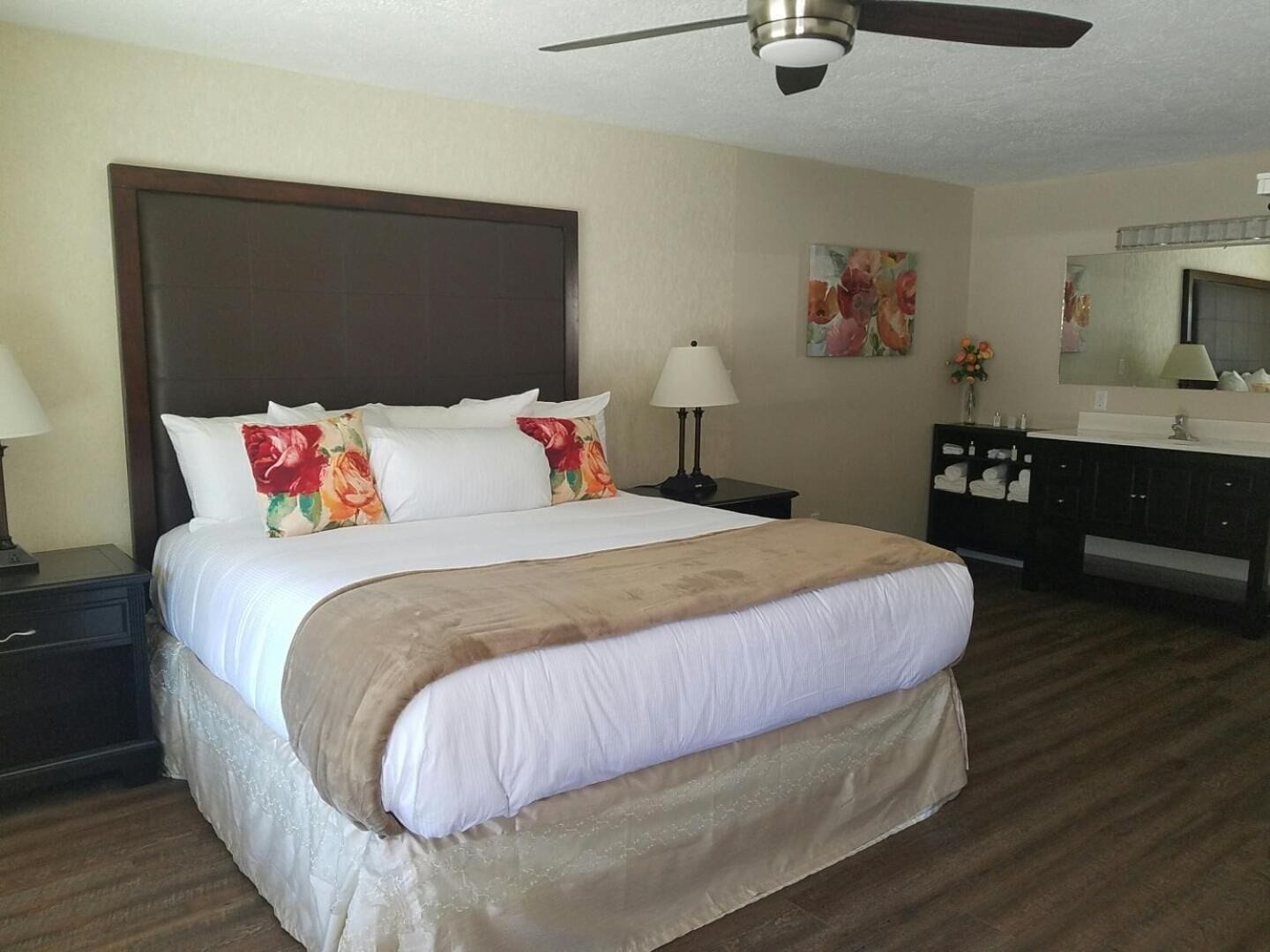 A hotel room with a large bed, a nightstand with a lamp, a dresser with a mirror, and a ceiling fan.