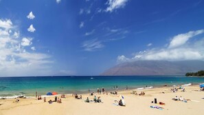 A beach with clear turquoise water, people sunbathing, and a mountain in the background.