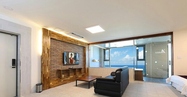 A modern hotel lobby with a brick wall, a flat-screen TV, a black leather sofa, and a glass door leading to a balcony with a view of the sea.