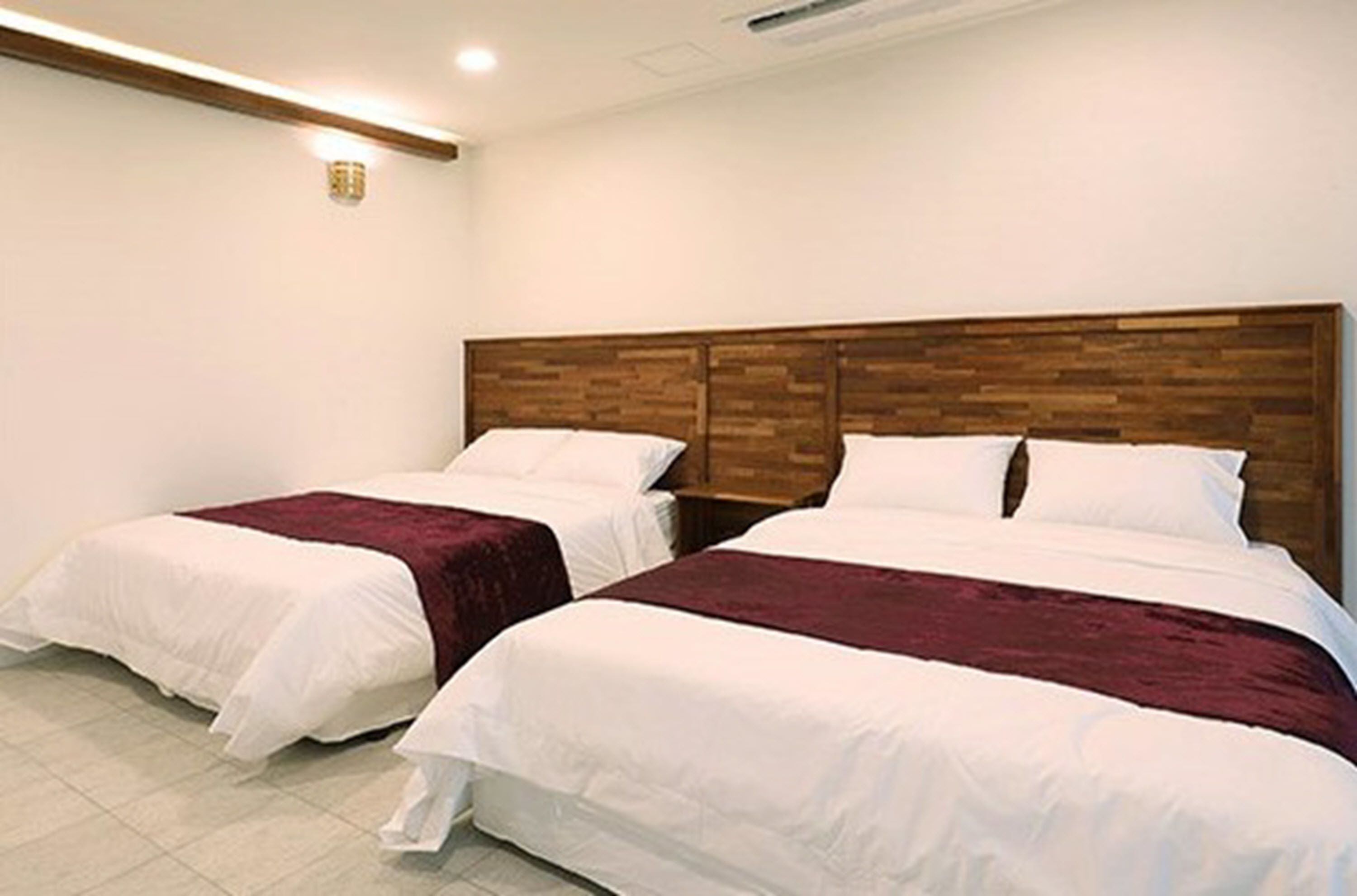 A hotel room with two single beds, each with white bedding and a maroon throw blanket. The headboard is made of wood, and there is a small wall-mounted light fixture above the bed.