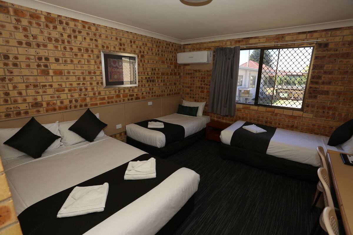 Une chambre d’hôtel avec des murs en briques, trois lits, une petite fenêtre avec des rideaux et un appareil de climatisation.