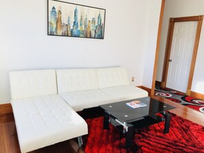 A white sofa, a glass coffee table, and a red rug in a room with a cityscape painting on the wall.