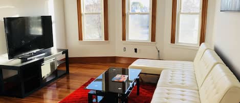 Un salon doté d’un téléviseur à écran plat, d’un canapé blanc, d’un tapis rouge et de trois fenêtres à cadre en bois.