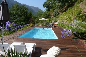 Piscina rectangular con cubierta de madera, rodeada de abundante follaje y flores moradas.