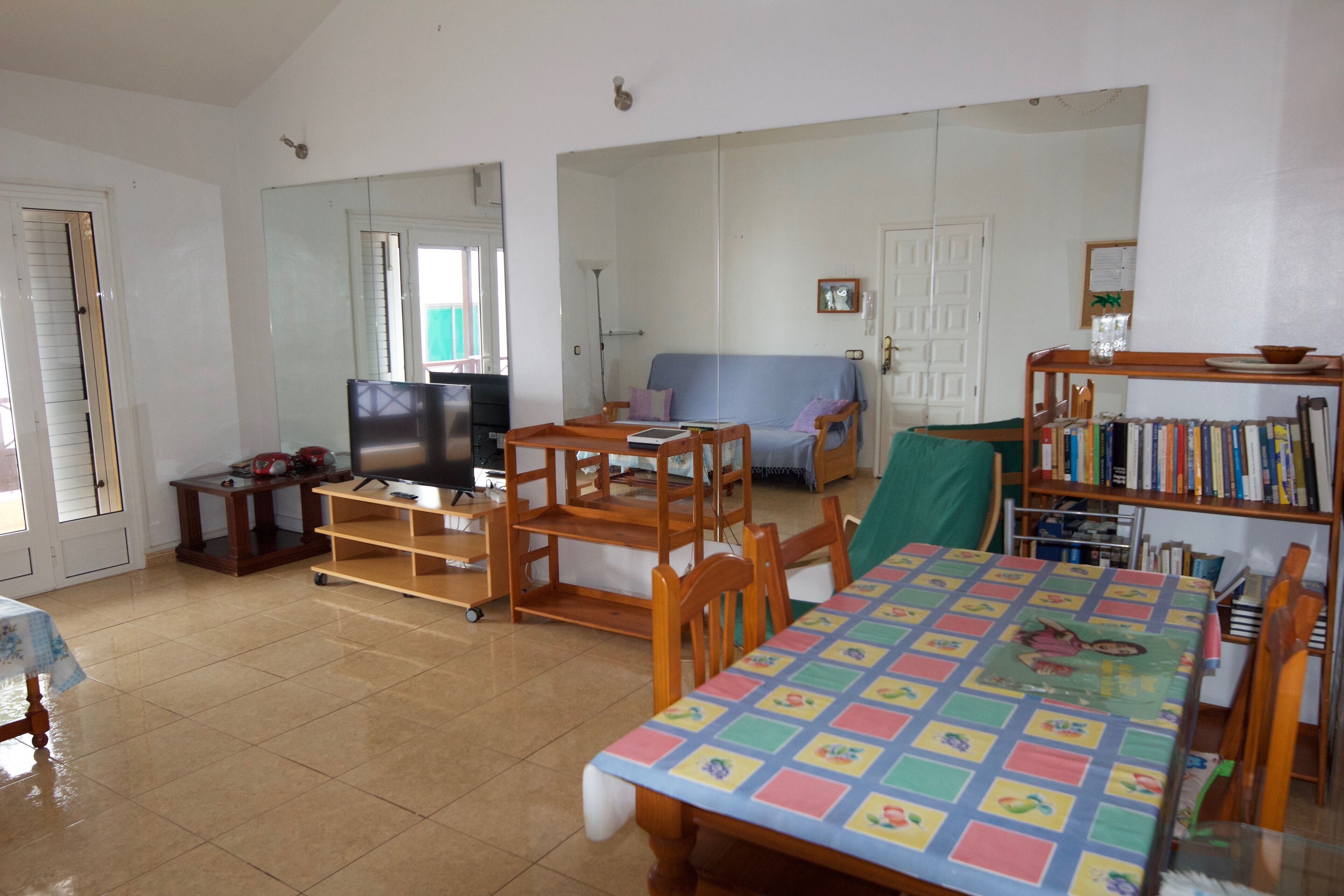 A spacious living room with a large TV, a sofa, a dining table with chairs, and a bookshelf.