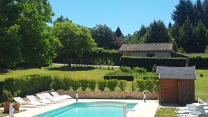 Una piscina con lettini, un piccolo deposito e una casa con tetto in tegole in un giardino curato.