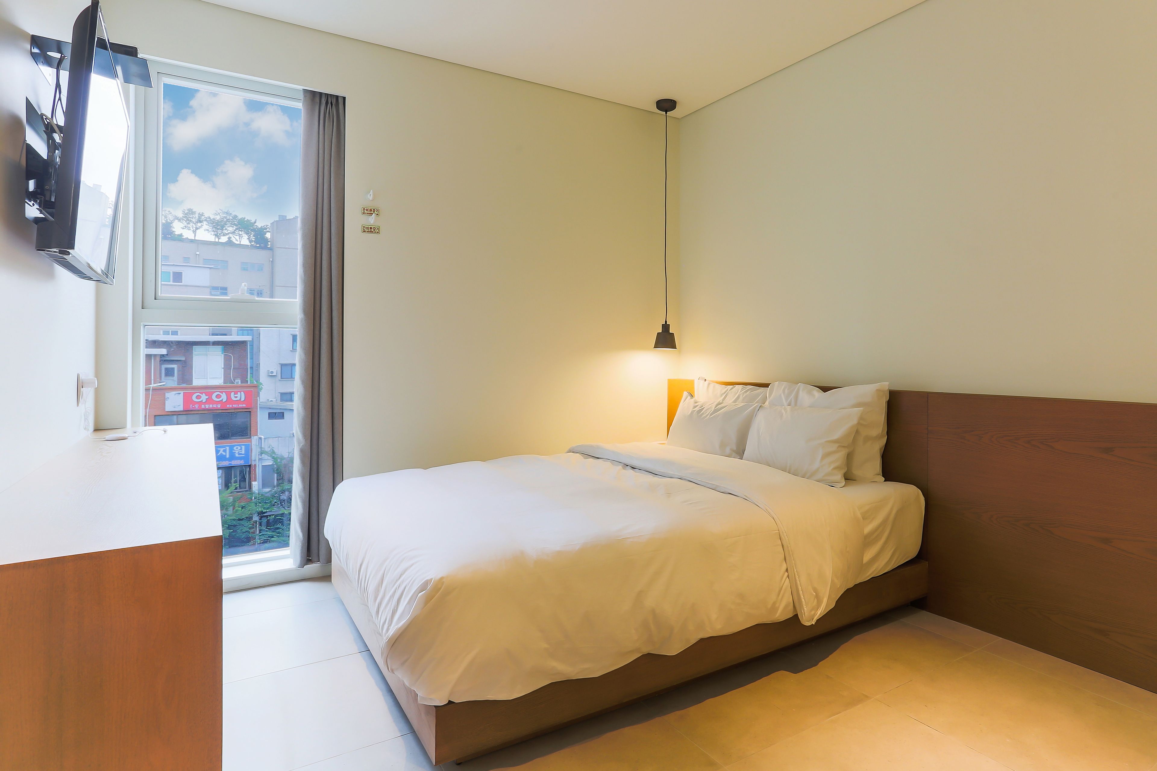 A bedroom with a bed, a bedside table, a lamp, and a window with a view of buildings.