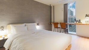 Quarto de hotel moderno com uma cama grande, uma mesa de trabalho com duas cadeiras, uma janela com vista para a cidade e uma luminária na parede.