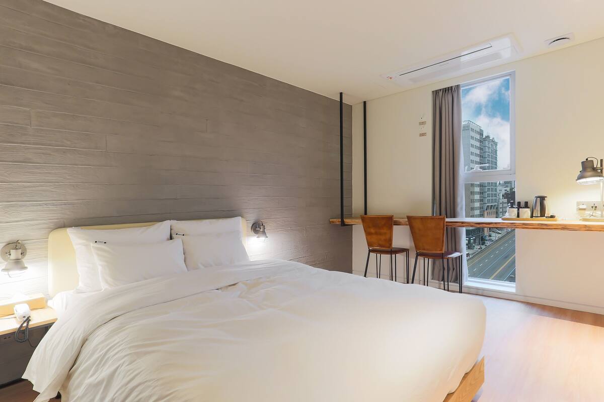 A modern hotel room with a large bed, a desk with two chairs, a window with a city view, and a wall-mounted lamp.