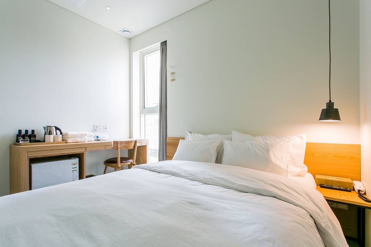 A hotel room with a bed, a desk with a computer, a chair, a small fridge, and a bedside table with a lamp and a phone.