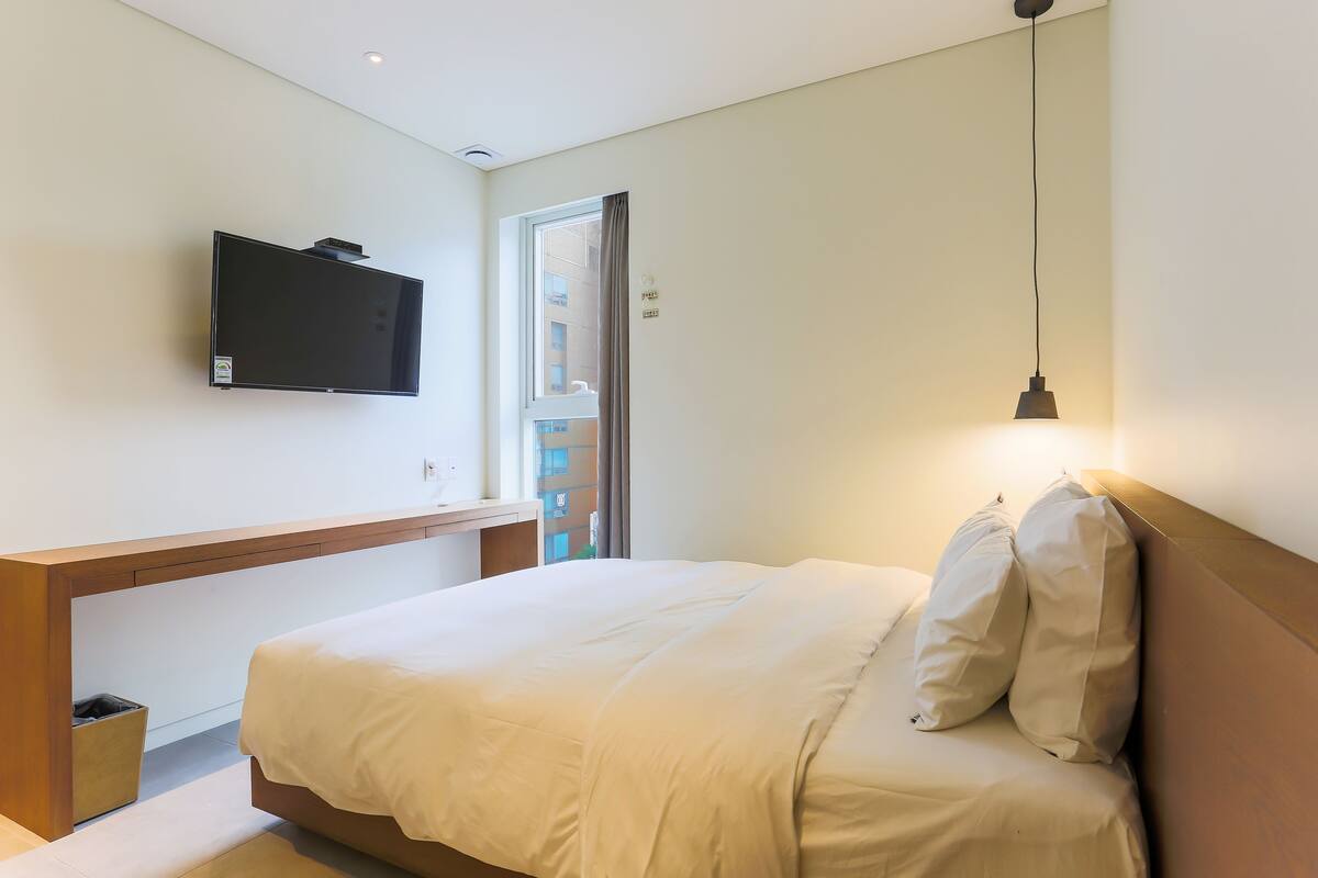 A modern hotel room with a bed, bedside table, TV, and a view of the outside.
