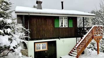 Eine zweistöckige Holzhütte mit einem mit Schnee bedeckten Dach, einem Schornstein und einer Holztreppe, die zu einer Terrasse führt.