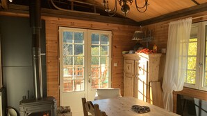 A cozy wooden cabin interior with a wood-burning stove, a dining table, and a view of the outside deck. - Dream cottage in the heart of nature: forest, river, cabin atmosphere & wood stove (Xonrupt-Longemer)