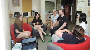 A group of people seated in a room with a laptop on a table, a clock on the wall, and a Coca-Cola refrigerator. - Hotel Lucky (Osaka)