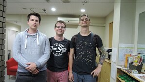 Three individuals standing in a room with a red chair and shelves with items on the right. - Hotel Lucky (Osaka)