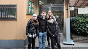 A group of people standing in front of a building with a glass door entrance, a signboard, and a metal awning. - Hotel Lucky (Osaka)