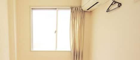Una habitación pequeña y limpia con suelo de tatami, una cama individual, una mesita de noche y una ventana con cortinas.