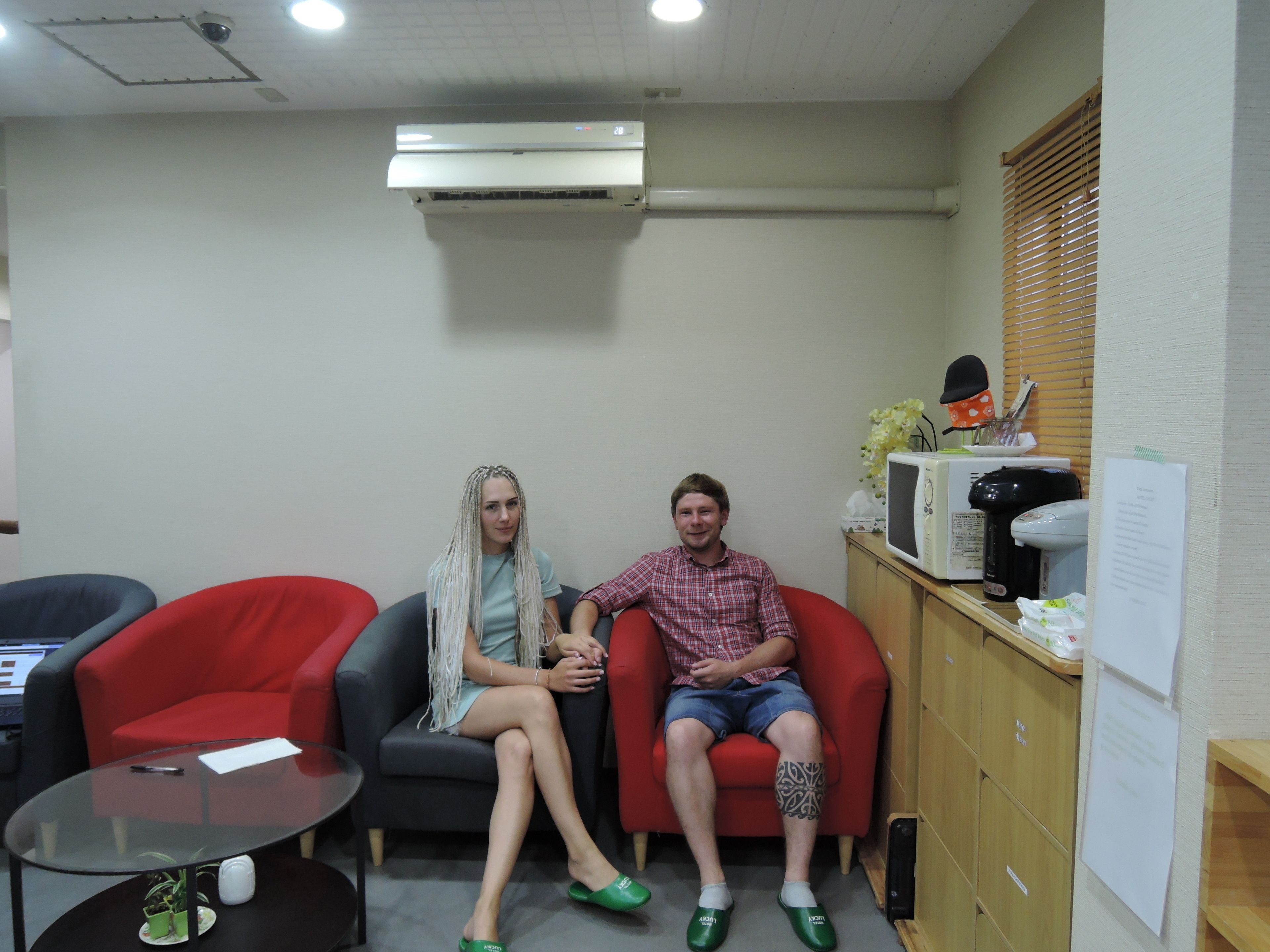 Two people seated on red chairs in a room with a glass table, a whiteboard, and a microwave on a wooden counter.