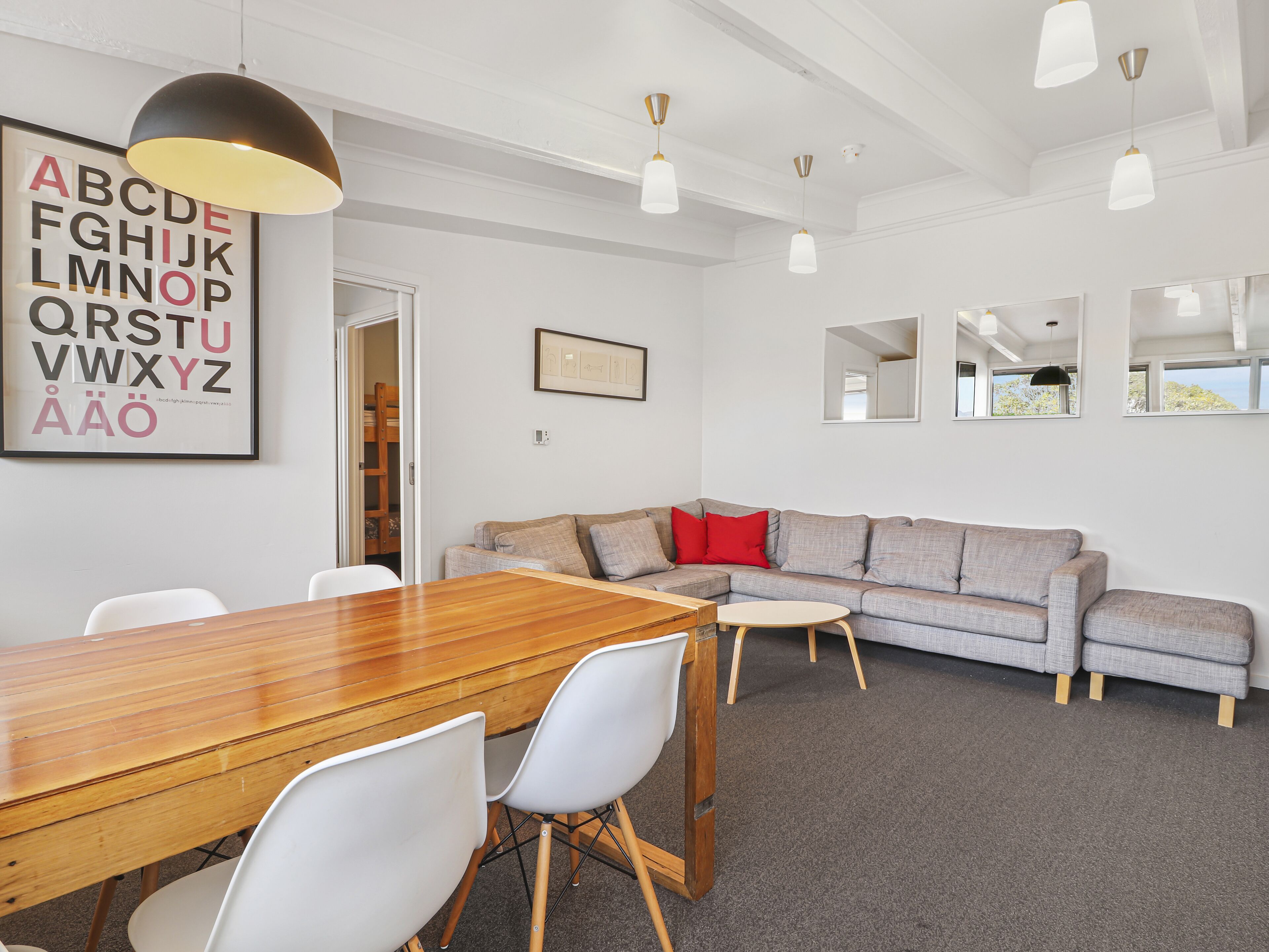 A modern living room with a wooden dining table, white chairs, a grey sofa with red cushions, and pendant lighting.