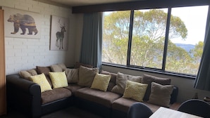 A living room with a sectional sofa, a dining table, and a large window with a view of trees and hills. - Snowflake 13 (Mount Buller)