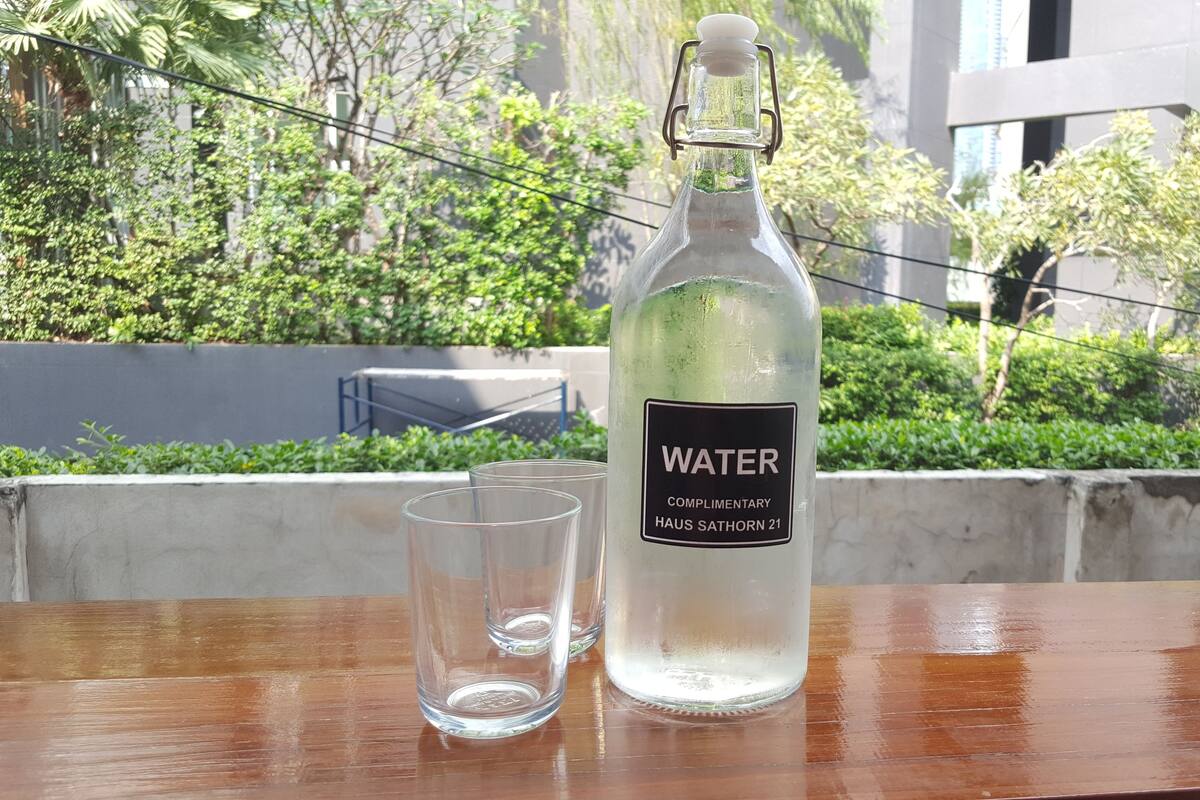 A bottle of water and a glass on a wooden table with a view of greenery and buildings in the background.