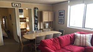 A cozy living room with a red couch, a dining table with chairs, a skateboard on the wall, and a window providing natural light. - Snowflake 14 (Mount Buller)