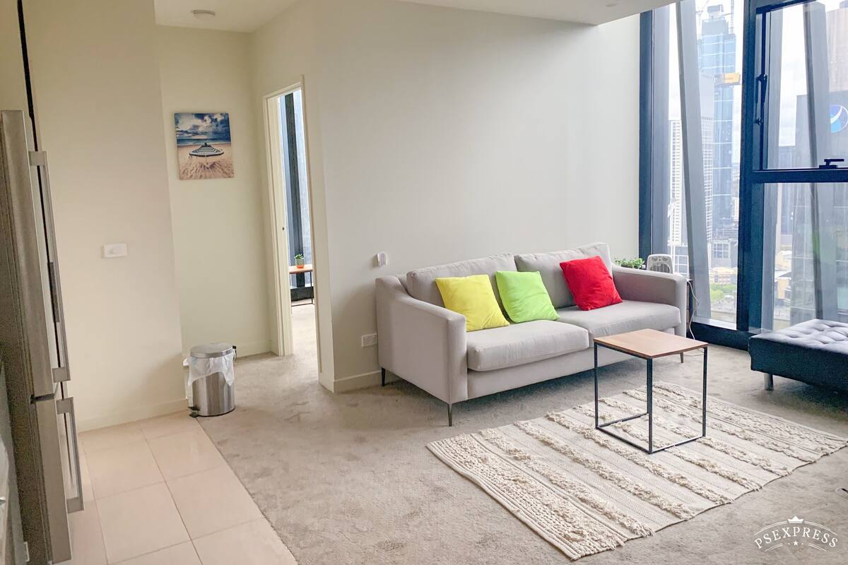 A modern living room with a grey sofa, colorful cushions, a small coffee table, and a large window offering a city view.