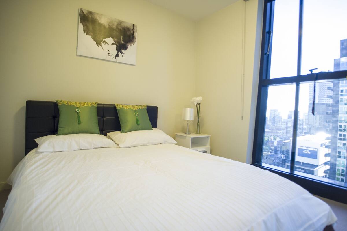 A hotel room with a large bed, a nightstand, a vase with flowers, and a city view through the window.